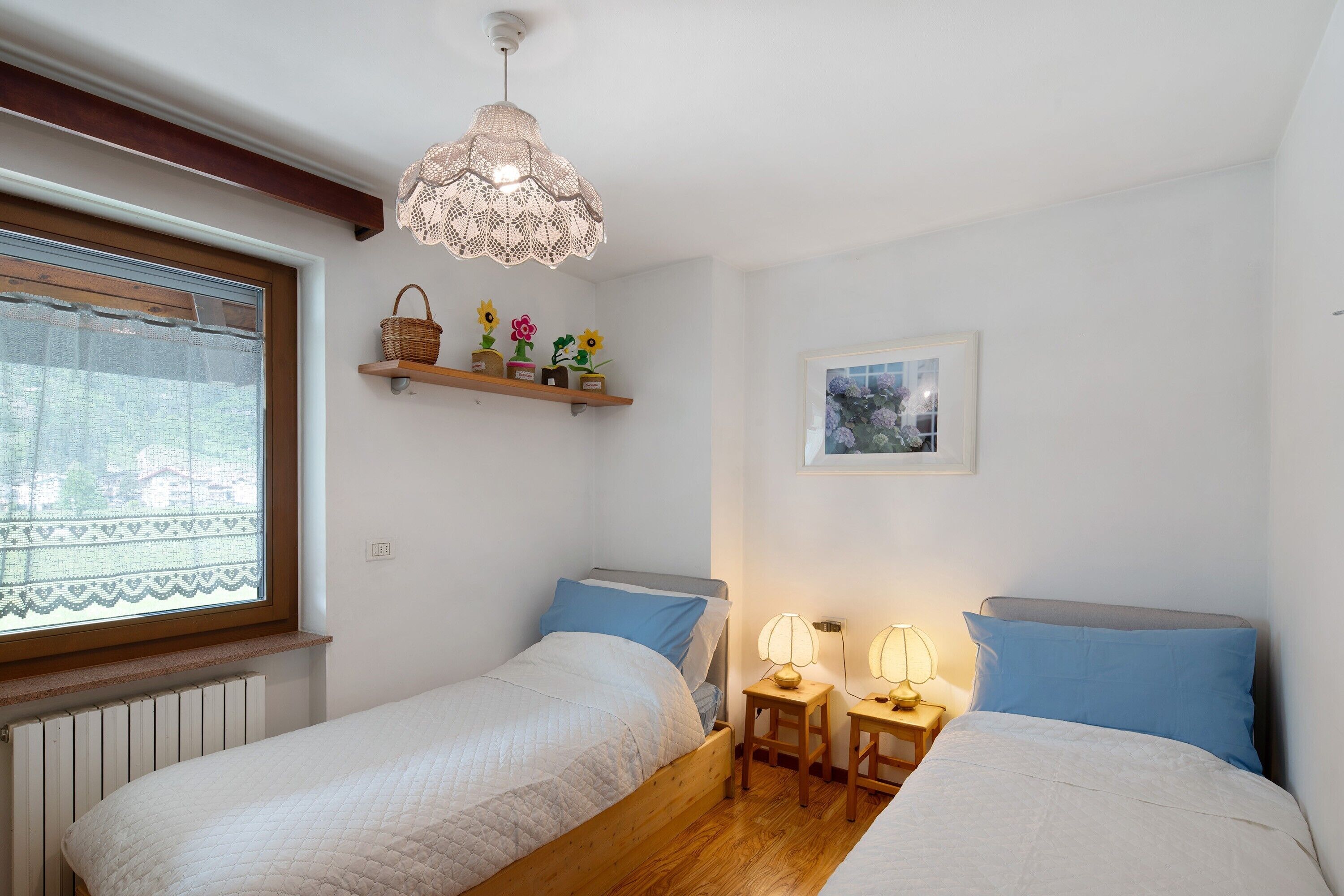 Ein Schlafzimmer mit zwei Betten, einem Fenster mit Spitzenvorhängen, einem Holzbalken an der Decke, einem an der Wand befestigten Regal mit Dekorationen und einer Hängelampe.