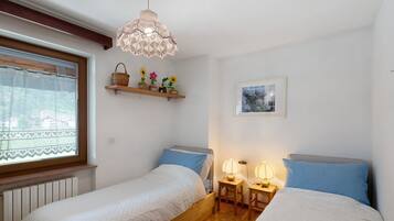 Ein Schlafzimmer mit zwei Betten, einem Fenster mit Spitzenvorhängen, einem Holzbalken an der Decke, einem an der Wand befestigten Regal mit Dekorationen und einer Hängelampe.