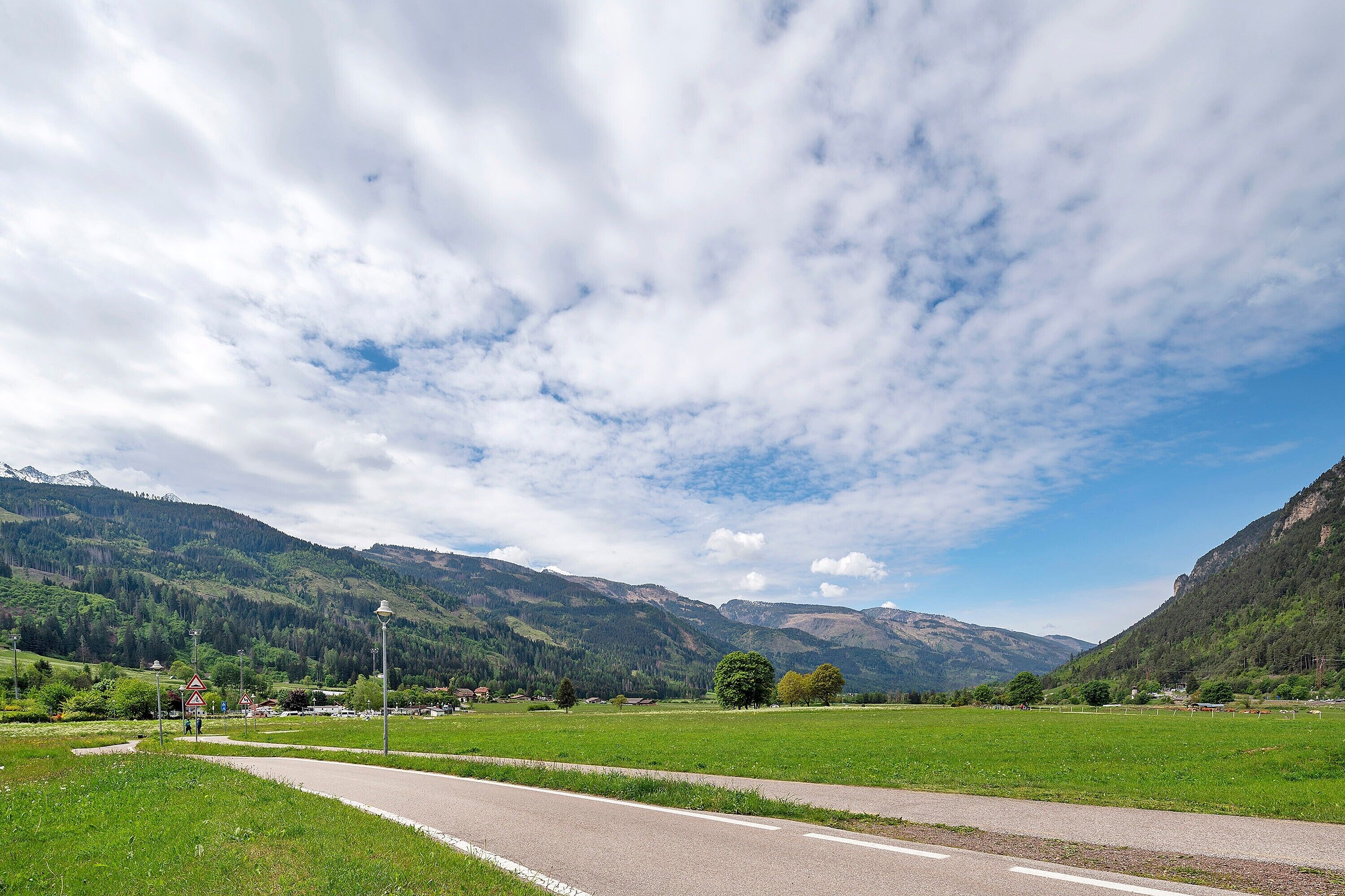 A rural road with a clear sky, green fields, and mountains in the background.
