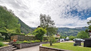 Un chemin pavé mène à une zone herbeuse bordée d’une clôture en bois, avec un petit arbre et une chaîne de montagnes en arrière-plan.