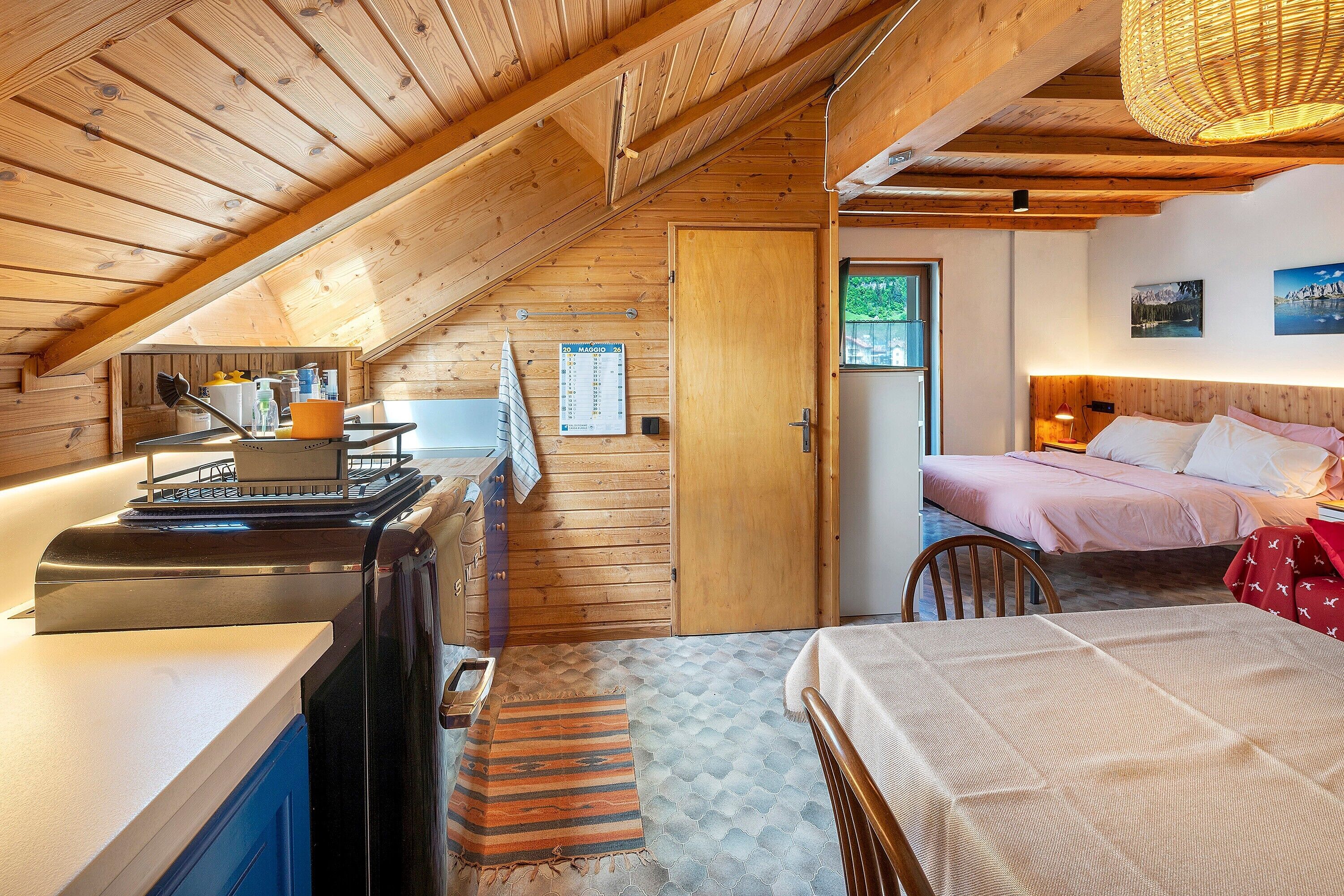 Een gezellig slaapkamer op zolder met een houten plafond, een bed, een klein tafeltje en een keukenhoek met een kookplaat.