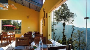 A dining area with tables set for two, wooden chairs, and a view of a mountainous landscape through large windows. - Mary's Chalet (Dalhousie)
