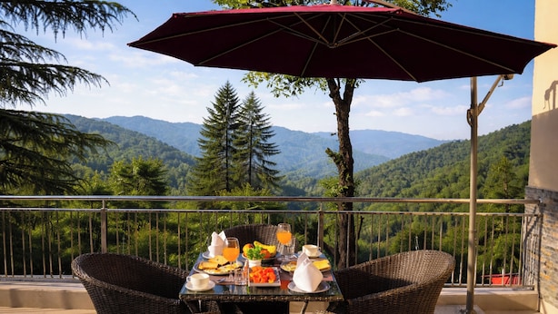 A balcony with wicker furniture, a table set with breakfast, and a view of mountains. - Mary's Chalet (Dalhousie)