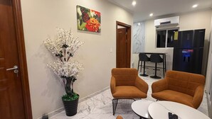 Ein Hotelzimmer mit Marmorboden, zwei orangefarbenen Sessel, einem kleinen weißen Tisch, einem Topf mit weißer Orchideenpflanze, einem Schmetterlingsbild und einer Küchenzeile mit Kühlschrank und Tisch.