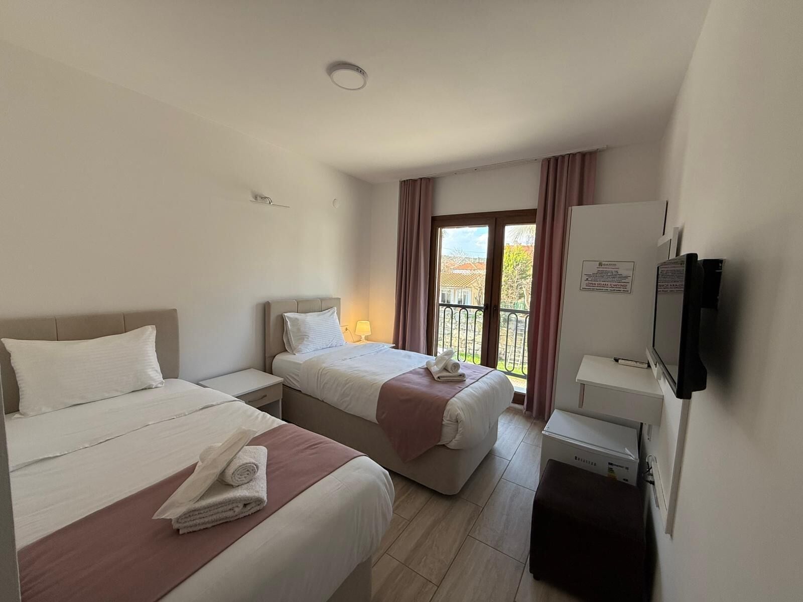 A hotel room with two beds, a TV, a desk, and a balcony with curtains.