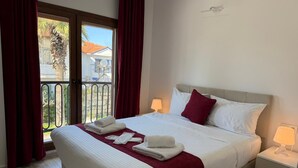 Een hotelkamer met een bed, twee bedlampen, een raam met gordijnen en uitzicht op een balkon.