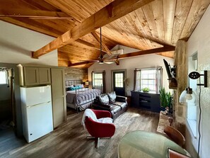 A spacious room with a high ceiling, wooden beams, a bed, a red chair, a dining table, and a refrigerator. - "Cheval Blanc" at Everwild Collective Cabins (Wimberley)