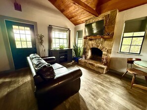 A living room with a stone fireplace, a leather sofa, a wooden ceiling, and a television mounted on the wall. - "Cheval Blanc" at Everwild Collective Cabins (Wimberley)
