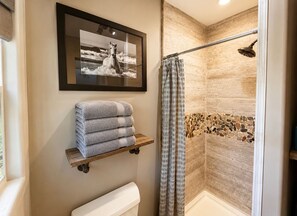 A bathroom with a shower, a framed picture, and a stack of towels on a shelf. - "The Surf Hut" at Everwild Collective Cabins (Wimberley)