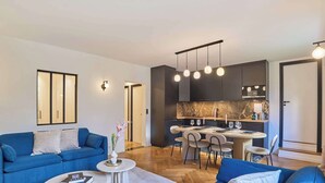 Un salon moderne avec un canapé bleu, un coin repas comprenant une table et des chaises, et une cuisine dotée de meubles sombres et d’un dosseret en marbre.