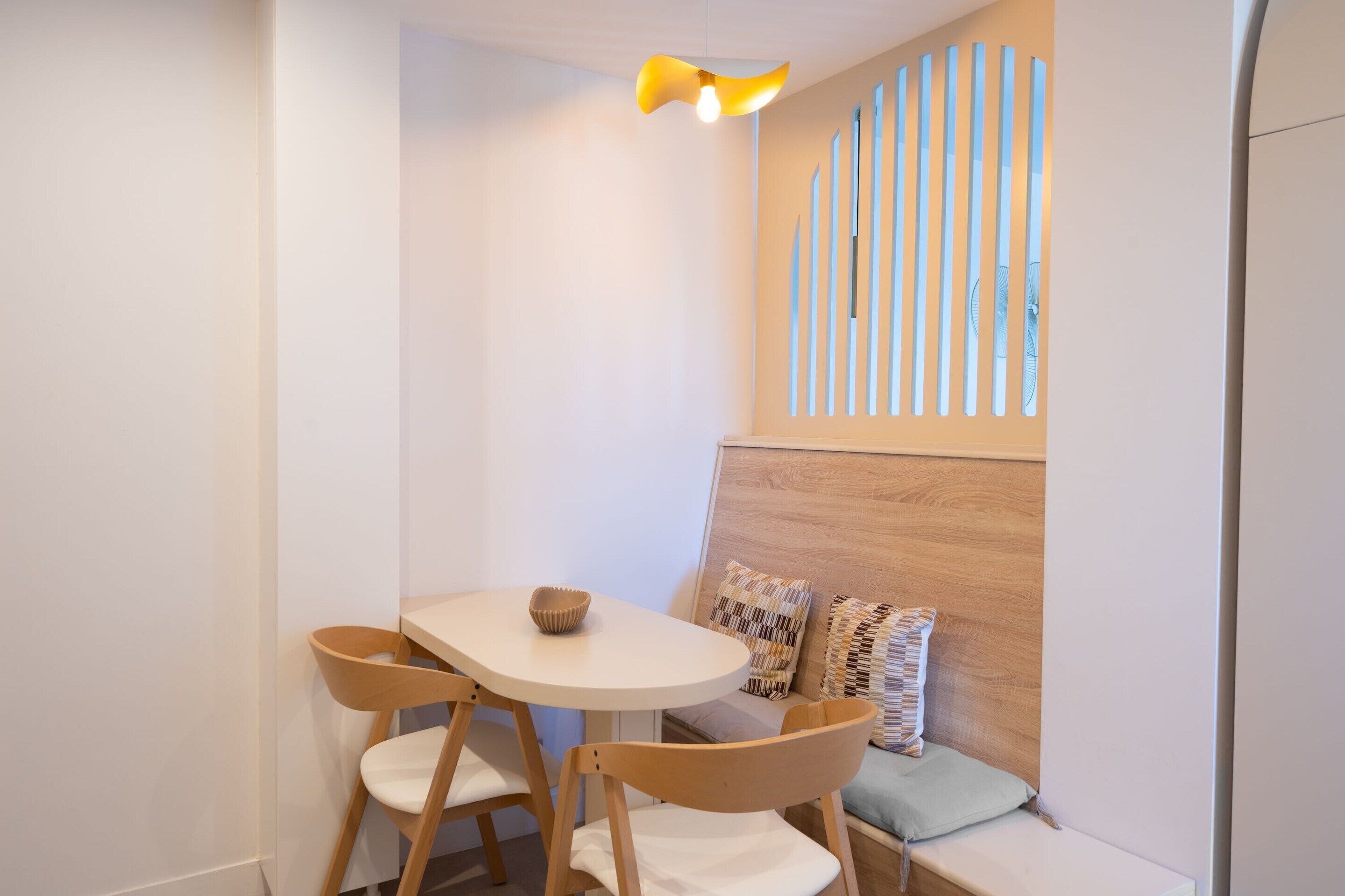Una habitación pequeña y acogedora con una mesa redonda, dos sillas de madera, un banco con cojines y una pared con listones verticales.