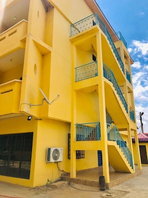 A multi-story yellow building with balconies, a visible air conditioning unit, and a staircase. - One Bedroom Apartment with Air Conditioning @ Royal Lanes Hotel, Achimota, Accra (Achimota, Accra.)