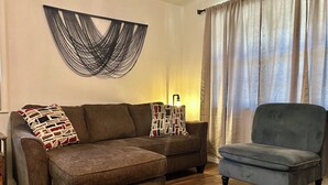 A living room with a brown sofa, a blue armchair, a patterned throw pillow, a wall-mounted abstract art piece, and a lamp on a side table.