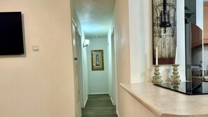 A hallway with a wooden floor, a light switch, a framed picture on the wall, and a kitchen area with cabinets and a countertop.