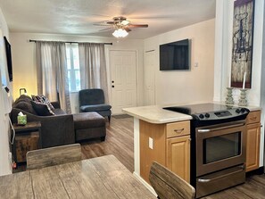 A compact kitchen with a stove, oven, and a small countertop. There is a dining table with chairs, a sofa, and a chair in the living area. A ceiling fan is visible, and curtains are on the windows.