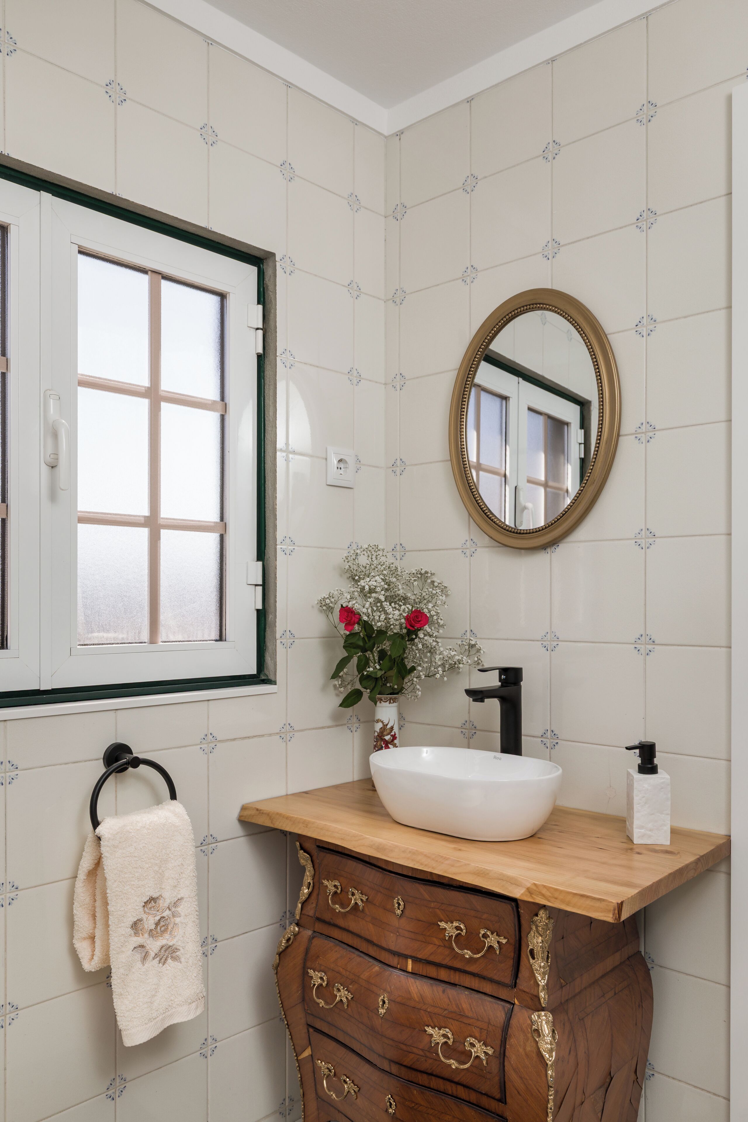Een badkamer met een witte wasbak, een houten wastafelmeubel, een spiegel, een raam en een handdoek aan een rek.
