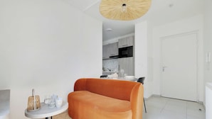 Ein modernes Wohnzimmer mit einer Couch, einem kleinen runden Tisch mit Gegenständen, einer Küche im Hintergrund und einer weißen Tür.