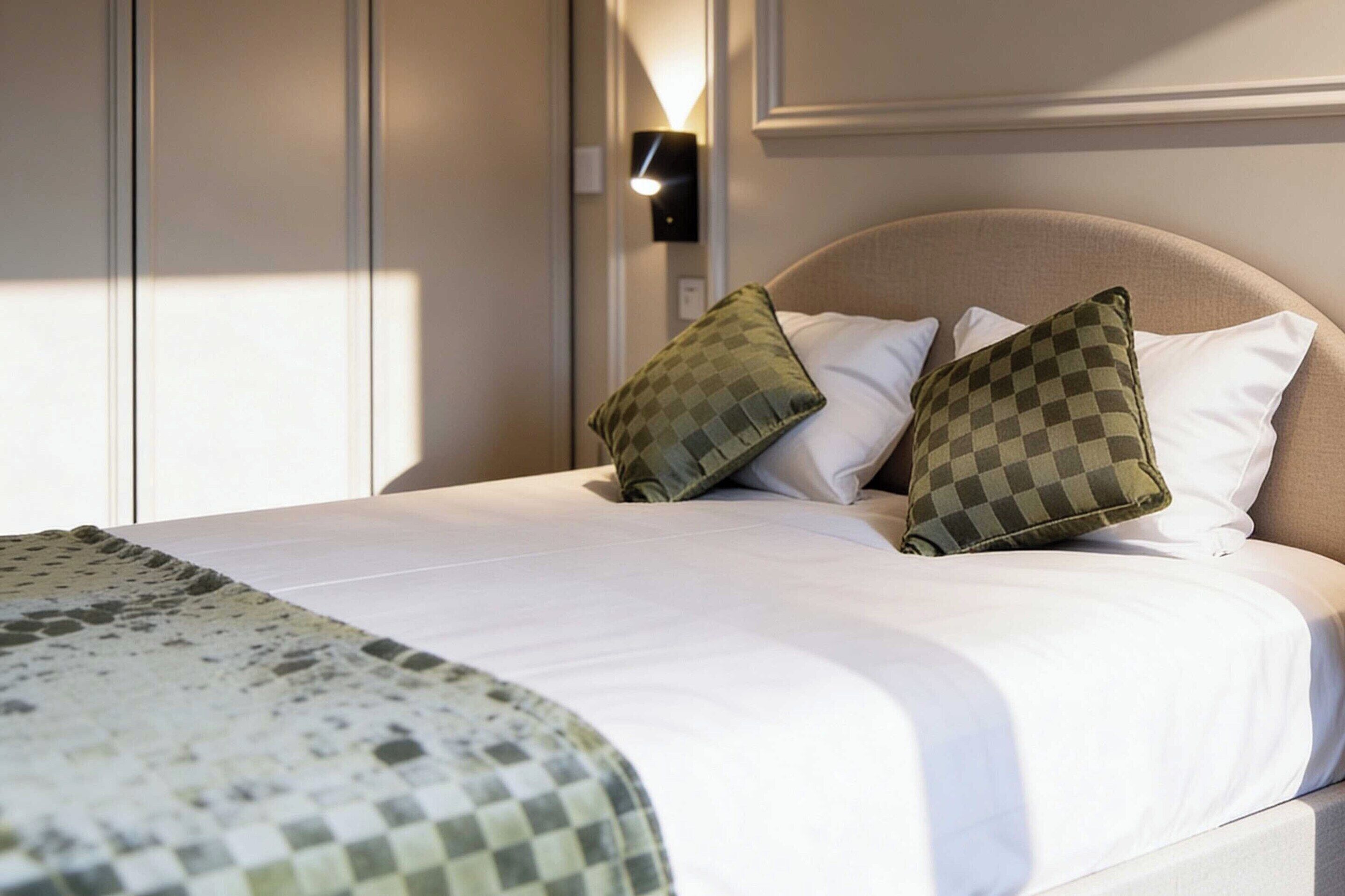 Een netjes opgemaakt bed met een geruit plaid, twee kussens en een wandlamp.