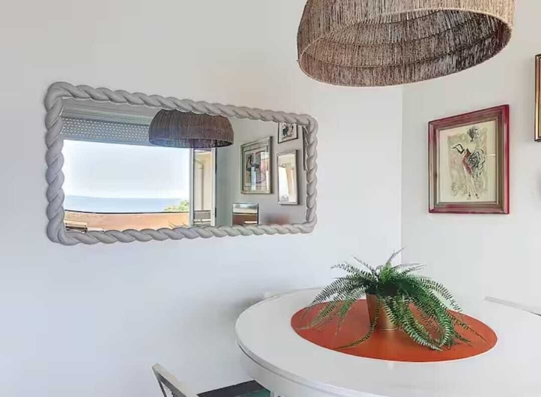 Una stanza con un tavolo rotondo bianco, una pianta in vaso, uno specchio con cornice in corda e un lampadario.