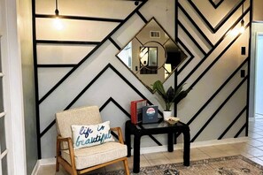 Ein Zimmer mit einer Wand mit geometrischem Muster, einem hölzernen Stuhl mit Kissen, einem kleinen Tisch mit Pflanze und einem Spiegel.