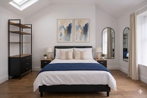 Une chambre à coucher comprenant un lit, deux tables de chevet, une table d’appoint, une bibliothèque, deux tableaux accrochés au mur, un miroir et une lucarne.
