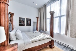 Un lit à baldaquin avec un cadre en bois, une grande fenêtre ornée de rideaux légers et deux œuvres d’art encadrées accrochées au mur.