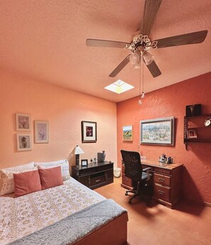 Ein Schlafzimmer mit Bett, Schreibtisch, Stuhl, Deckenventilator und Bildern an den Wänden.