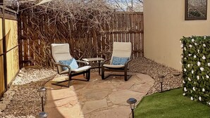 Un patio con dos sillas, una mesa y un pequeño jardín con seto.