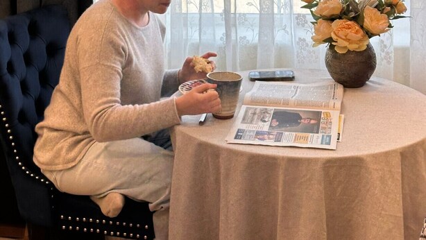 Una persona sentada a una mesa redonda con un libro y una taza, cerca de una ventana con cortinas traslúcidas y un jarrón de flores.