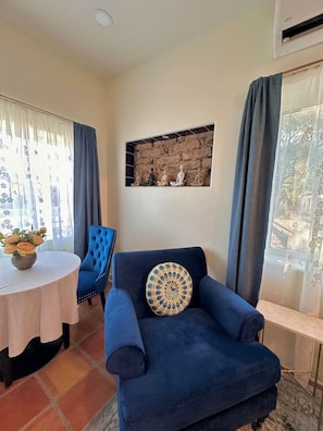 A room with a blue armchair, a round table with a vase of flowers, a wall-mounted shelf with sculptures, and a window with curtains.