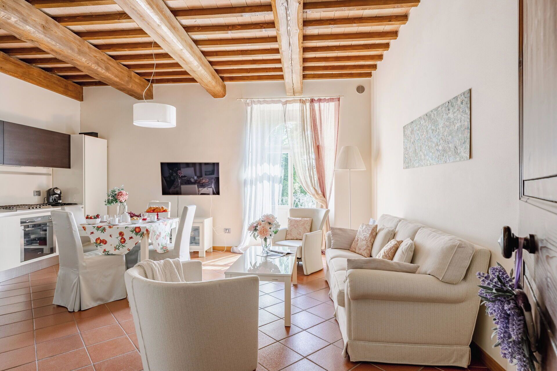 A living room with a sofa, chairs, a TV, a kitchen area, and a dining table with floral tablecloths.