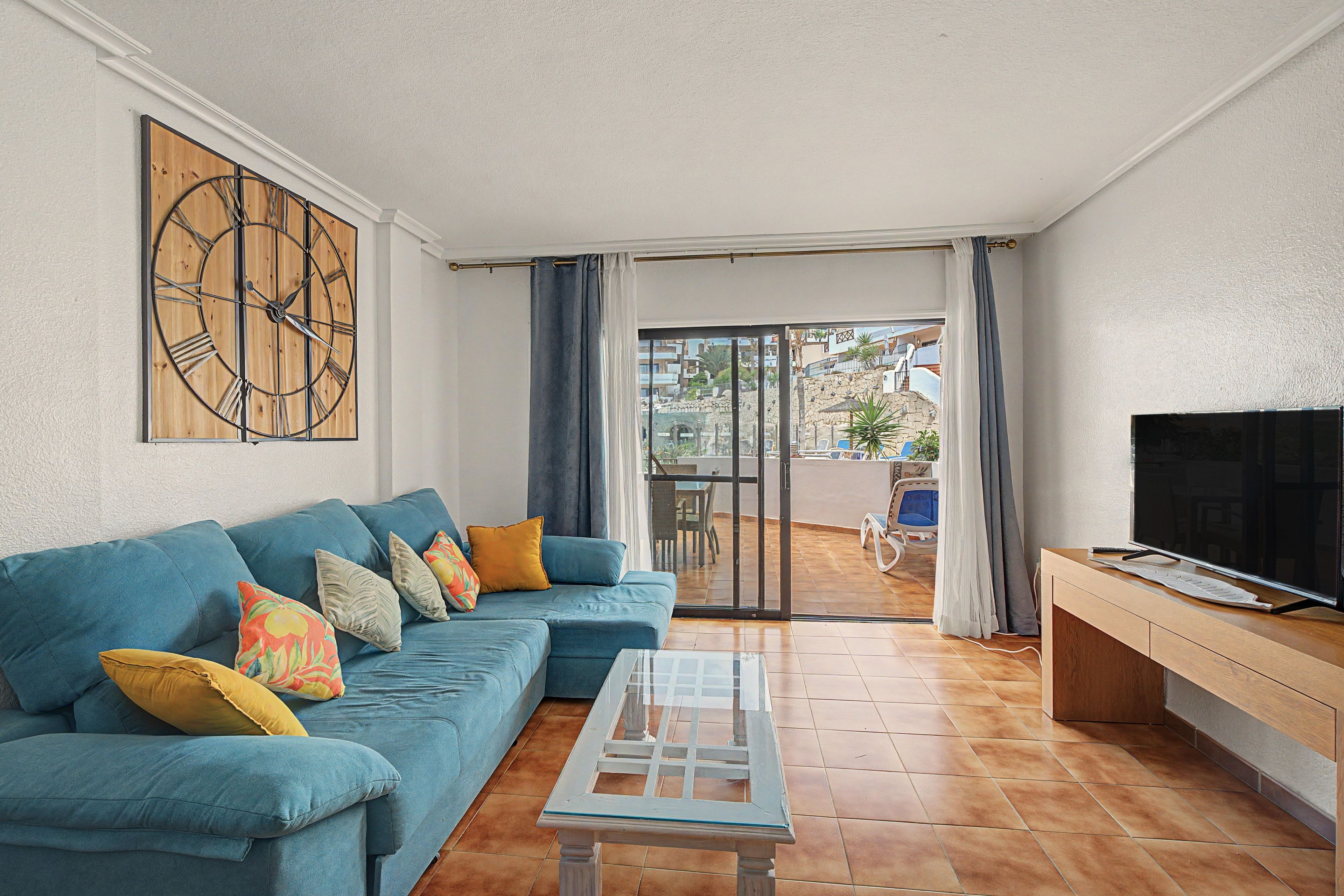 Sala de estar con un sofá azul, una mesa de centro, un reloj de pared grande, un televisor y vistas al exterior.