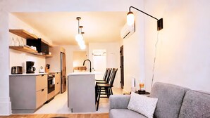 Een moderne woonkamer met een grijze bank, een salontafel met boeken, een eethoek met stoelen en een keuken met apparatuur.