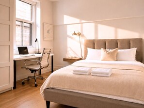 Une chambre à coucher comprenant un lit, un bureau avec un ordinateur portable, une chaise, une table de chevet avec une lampe et un luminaire fixé au mur.