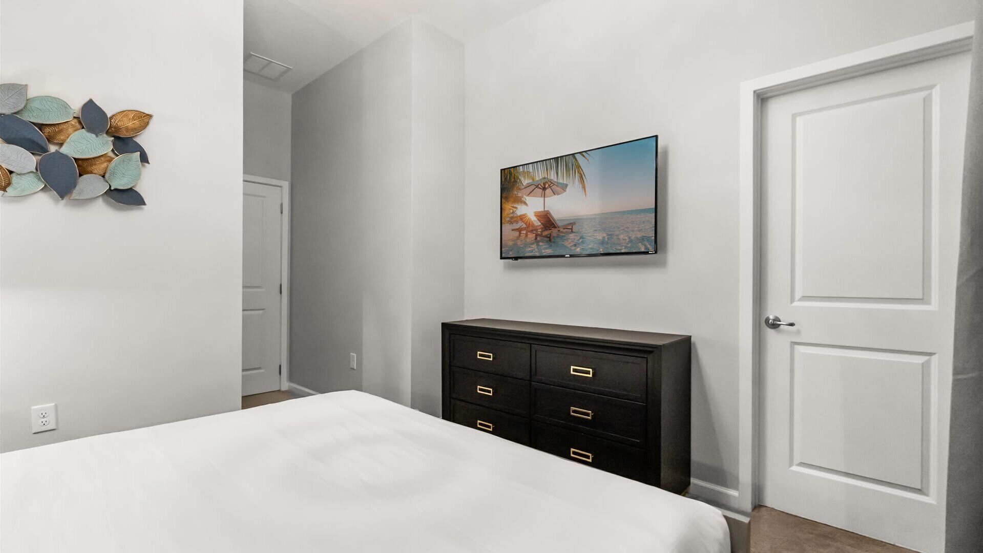 Un dormitorio con cama, cómoda, televisor montado en la pared y una puerta.