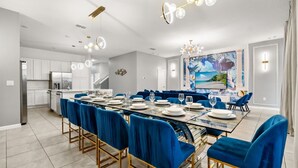 A modern dining room with a glass table, blue velvet chairs, and a large painting of a beach scene. - Upstay Bella Vida Retreat w Pool, Cinema and Games (Kissimmee)