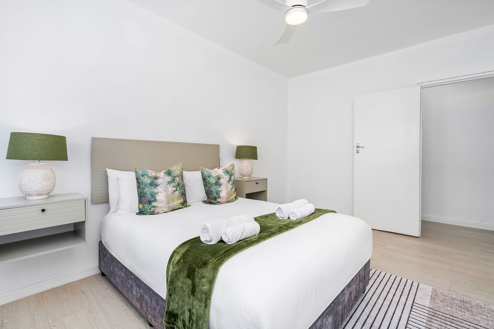 A neatly arranged hotel room with a bed, two bedside tables, a nightstand, a green throw blanket, and a ceiling fan.