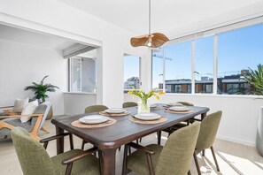 Un coin repas avec une table en bois dressée pour six personnes, entourée de chaises à revêtement vert, un luminaire suspendu et une vue sur les bâtiments à travers de grandes fenêtres.
