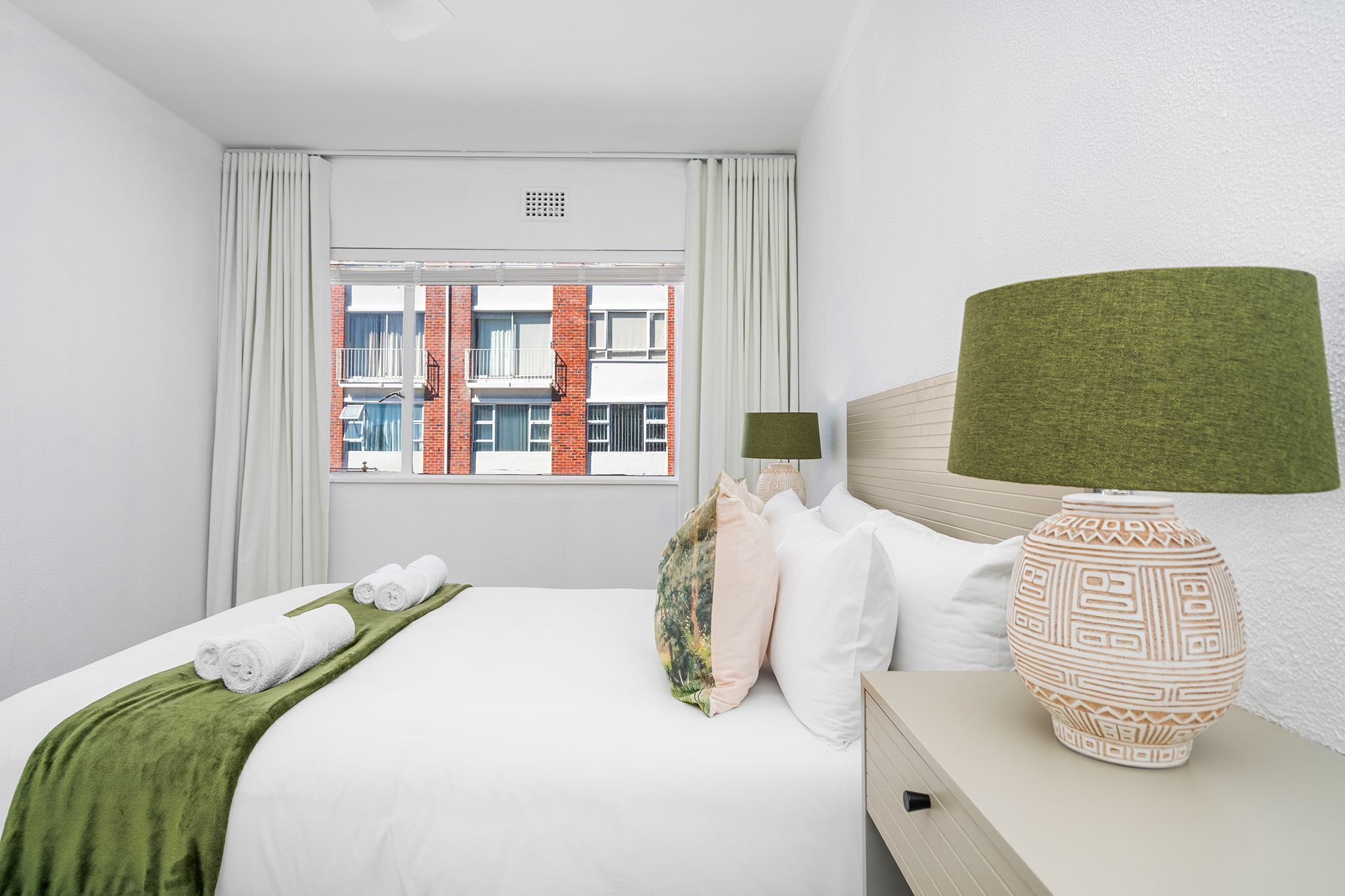 Une chambre d’hôtel avec un lit, des oreillers, un plaid vert, une lampe et une fenêtre donnant sur des immeubles.