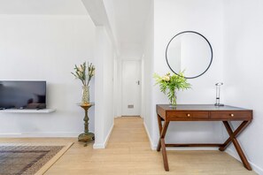 Une pièce avec un téléviseur posé sur une étagère, un bureau agrémenté d’un vase de fleurs et un miroir rond accroché au mur.
