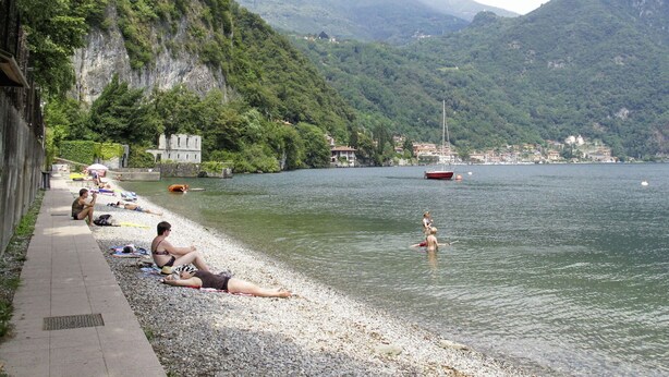 Vista del lago con una playa de guijarros, una fila de sombrillas y un velero a lo lejos.