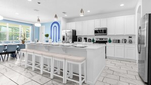 A modern kitchen with white cabinetry, a central island with bar stools, stainless steel appliances, and pendant lighting. - Upstay Family Resort Mansion- Pool, Games, Theater (Kissimmee)