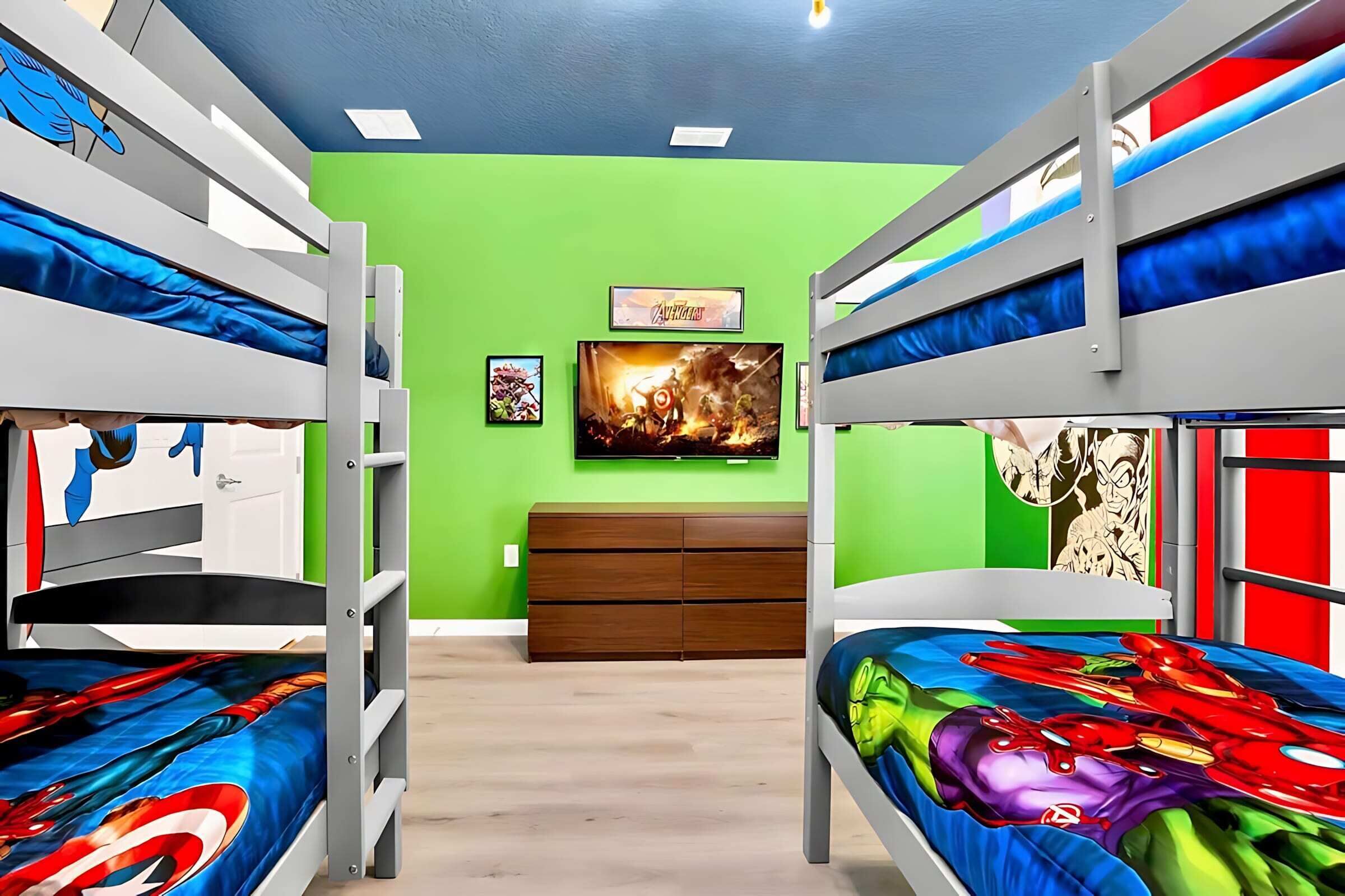 Une chambre avec un lit superposé, de la literie sur le thème des super-héros, un téléviseur fixé au mur et une commode en bois.