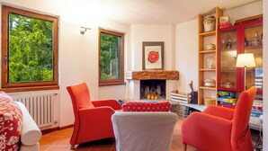 Uma sala de estar aconchegante com lareira, poltronas vermelhas, um banquinho listrado e uma estante com diversos objetos.