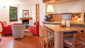 Uma sala de estar aconchegante com lareira, poltronas vermelhas, uma mesa de centro de madeira e uma área de cozinha com armários e uma luminária.