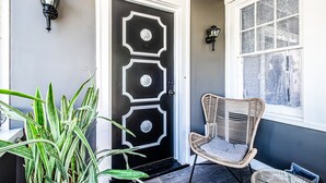 Une porte noire ornée de motifs décoratifs, une chaise en osier, une plante en pot et une fenêtre avec des rideaux à motifs.