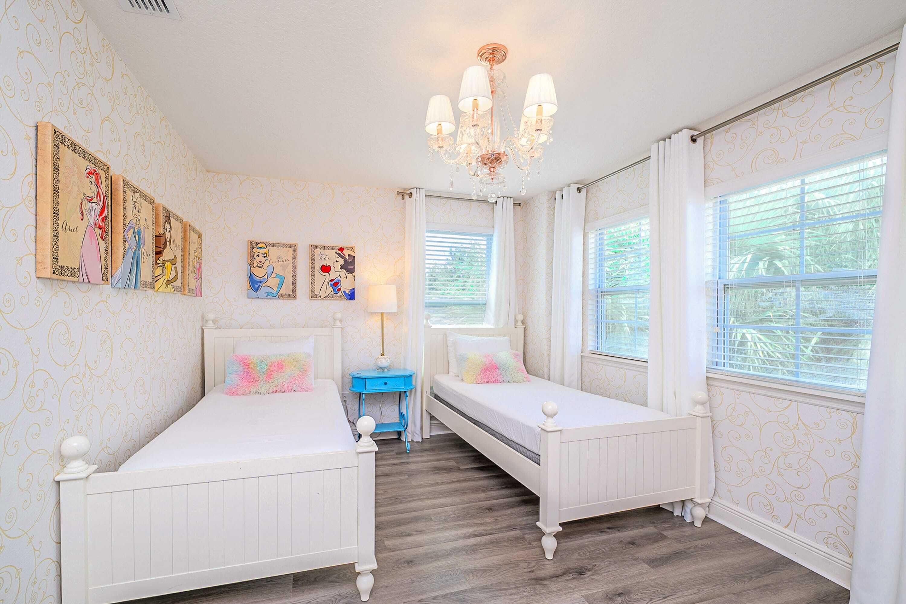 A bedroom with two white beds, a chandelier, and a window with blinds.