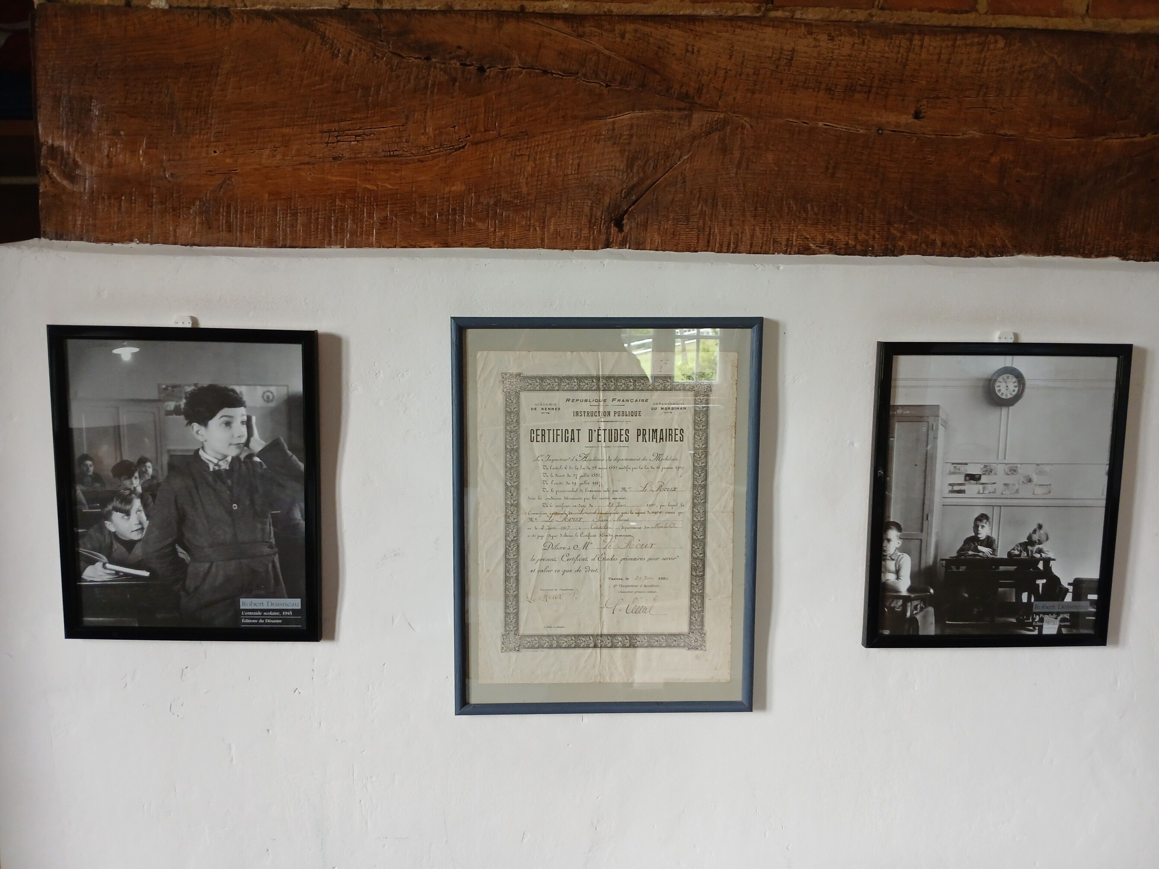 Drie ingelijste objecten aan een muur: een zwart-wit foto van een klaslokaal, een diploma basisonderwijs, en nog een zwart-wit foto van een klaslokaal.