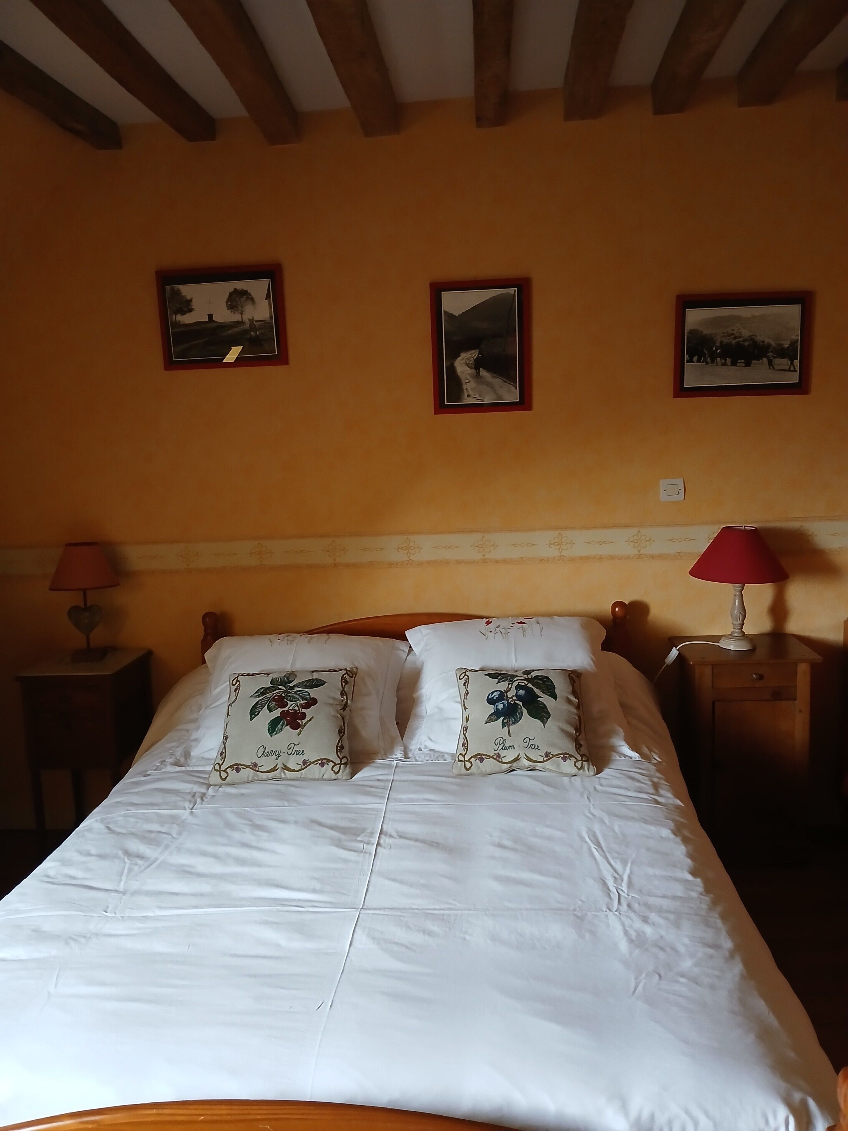 Un lit avec une literie blanche et des coussins à motifs floraux, une tête de lit en bois et une table de chevet avec une lampe rouge.