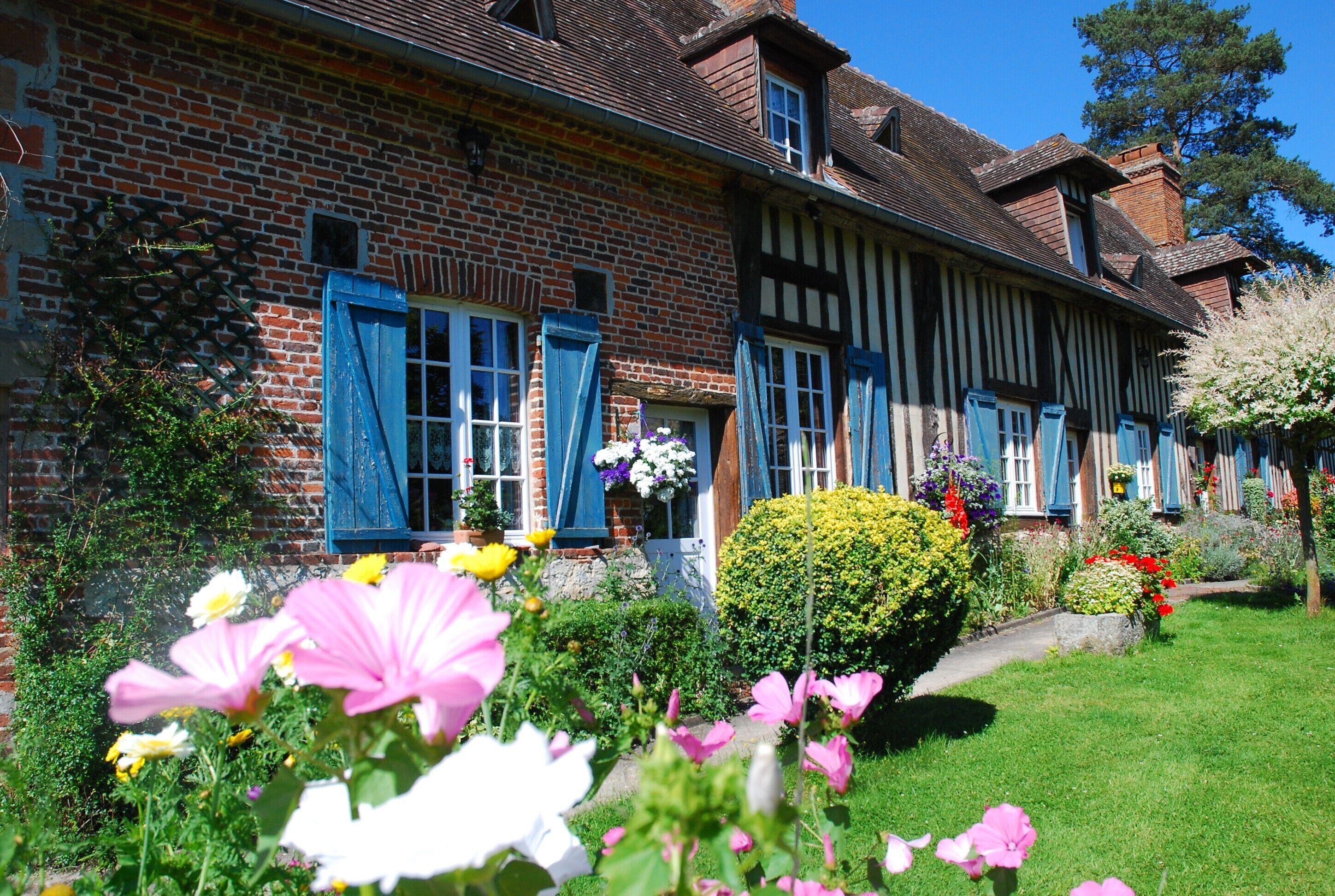 A traditional half-timbered house with a brick facade, multiple windows with shutters, and a well-maintained garden with various flowers and shrubs.